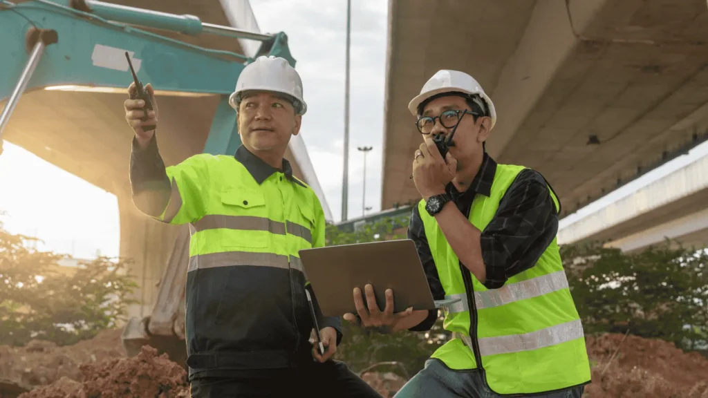 Professional insurance adjusters or contractors wearing safety vests and hard hats, inspecting a job site and using a laptop to document damage details.